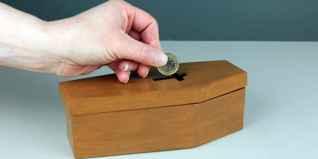 hand putting a pound coin into a small coffin money box