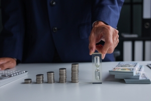 Hand Holding Small Hourglass Near Stacks of Coins and Cash